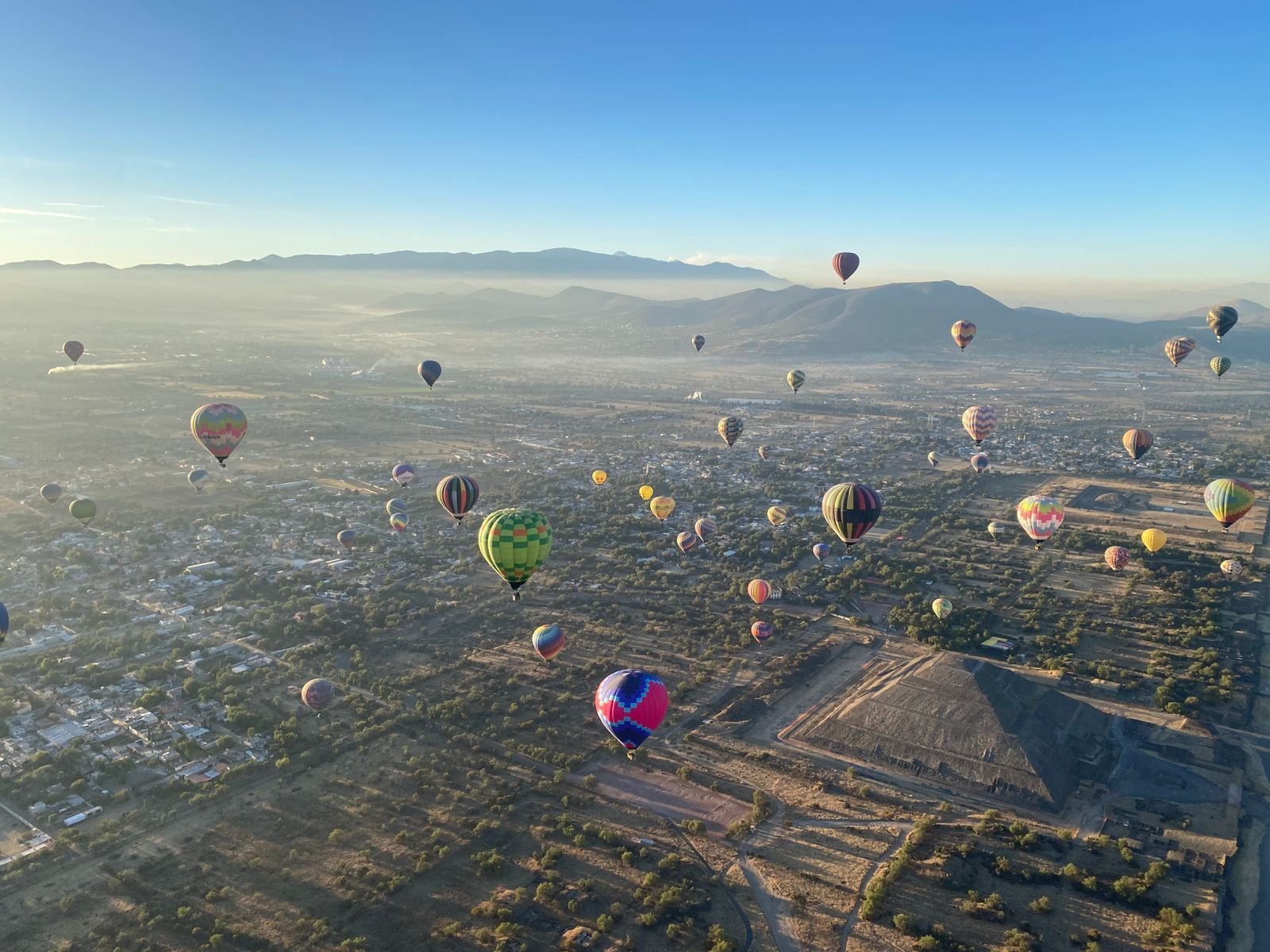 Decenas de globos aerostáticos coloridos flotando sobre un valle con montañas y una estructura de pirámide visible debajo en una mañana clara