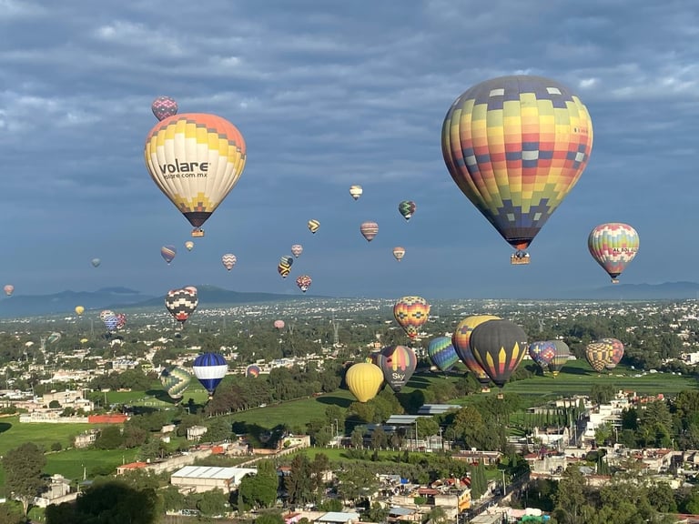 Múltiples globos aerostáticos flotando sobre un paisaje suburbano con campos verdes y casas en un día nublado