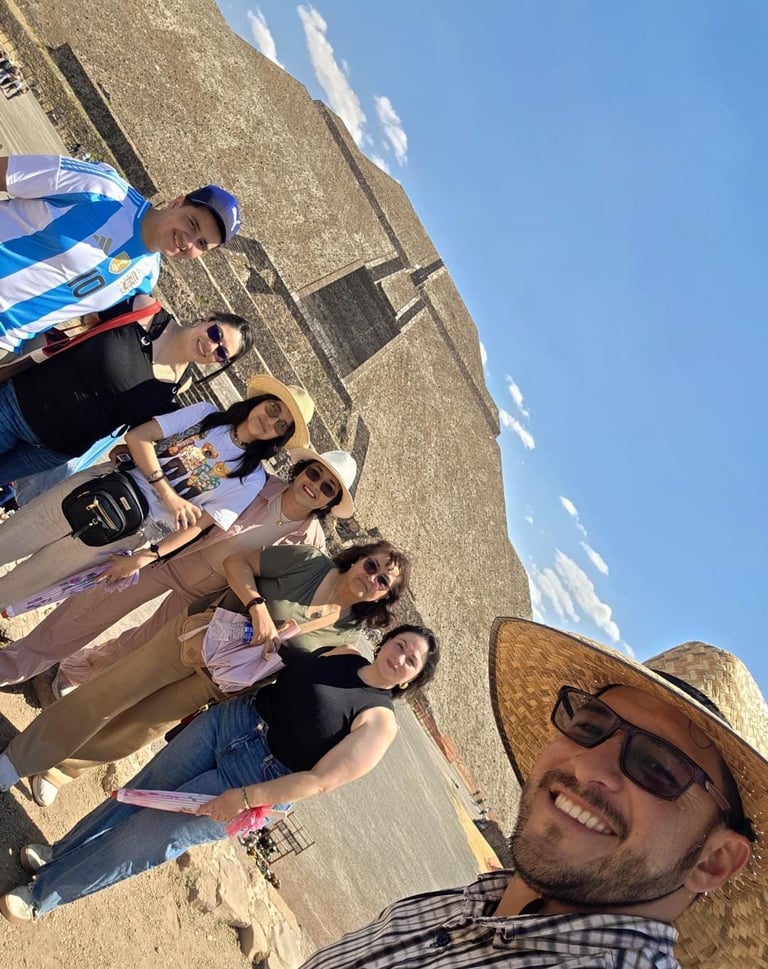Group of people standing on sandy beach with clear blue sky, man in foreground wearing straw hat and sunglasses taking selfie