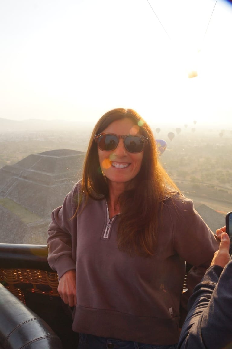 Woman wearing sunglasses smiling in a hot air balloon basket with landscape view below at sunrise