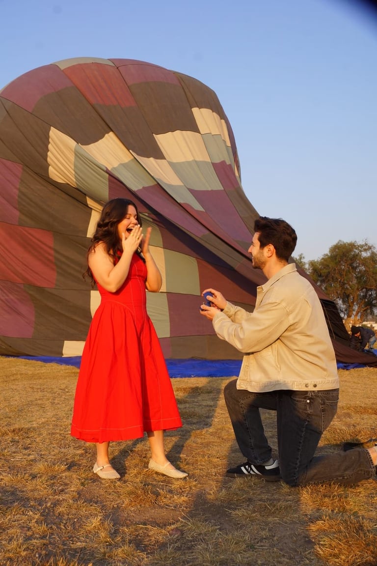 Man proposing to woman in red dress with hot air balloon in background during daytime