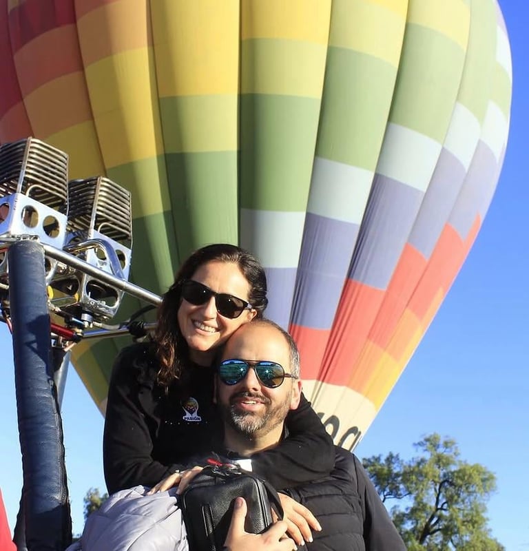 Dos personas sonriendo a la cámara con globo aerostático colorido y cielo azul de fondo