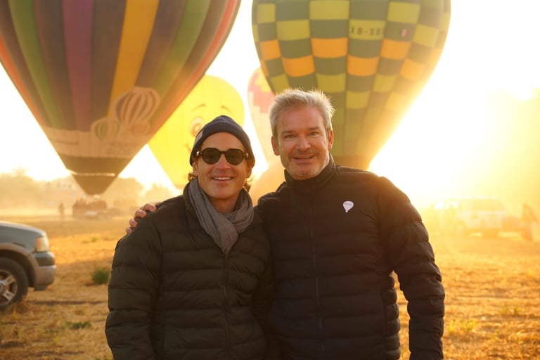 Two men standing together at sunrise with hot air balloons in the background during early morning flight preparation
