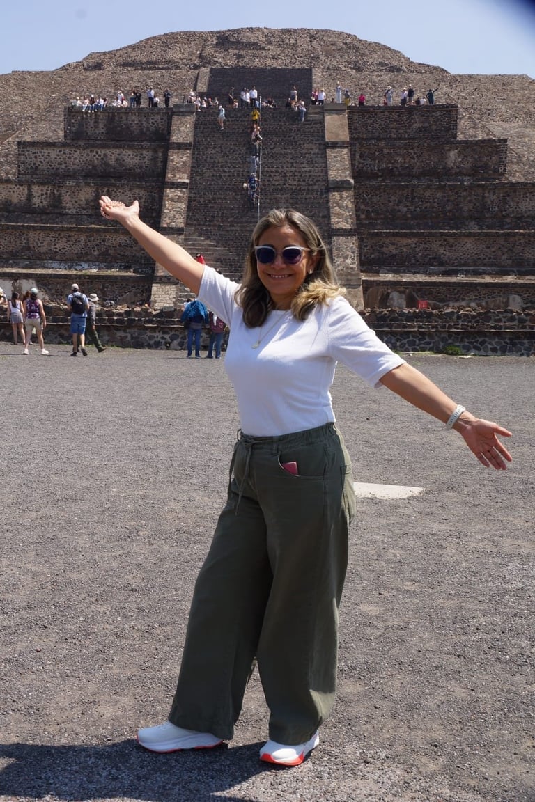 Woman posing with arms outstretched in front of an ancient Mesoamerican pyramid with stone structures visible in the background