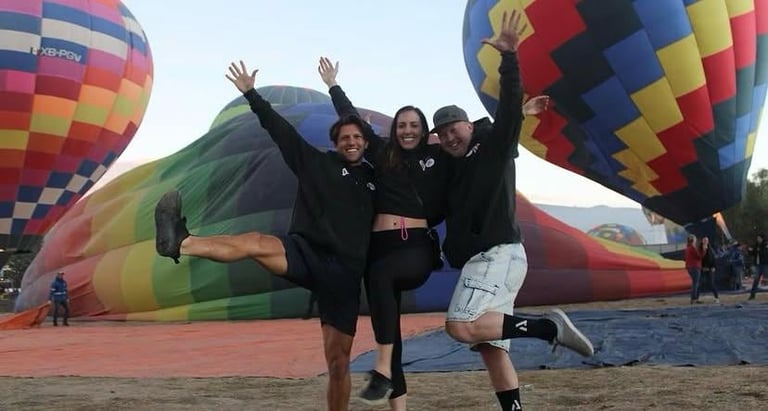 Tres personas saltando con brazos levantados en celebración frente a globos aerostáticos coloridos en el suelo