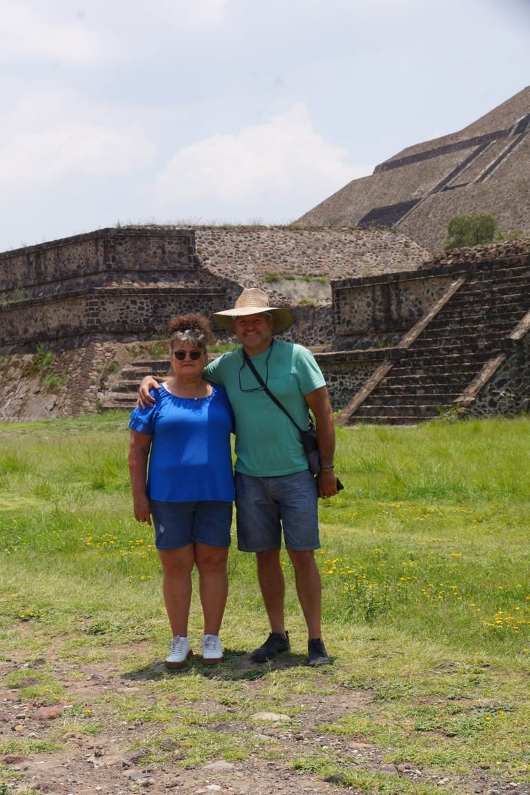 Two tourists posing in front of ancient stone pyramid ruins with stepped architecture and green grass field