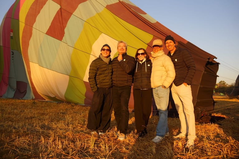 Six people standing in front of a colorful hot air balloon on a grassy field at sunset