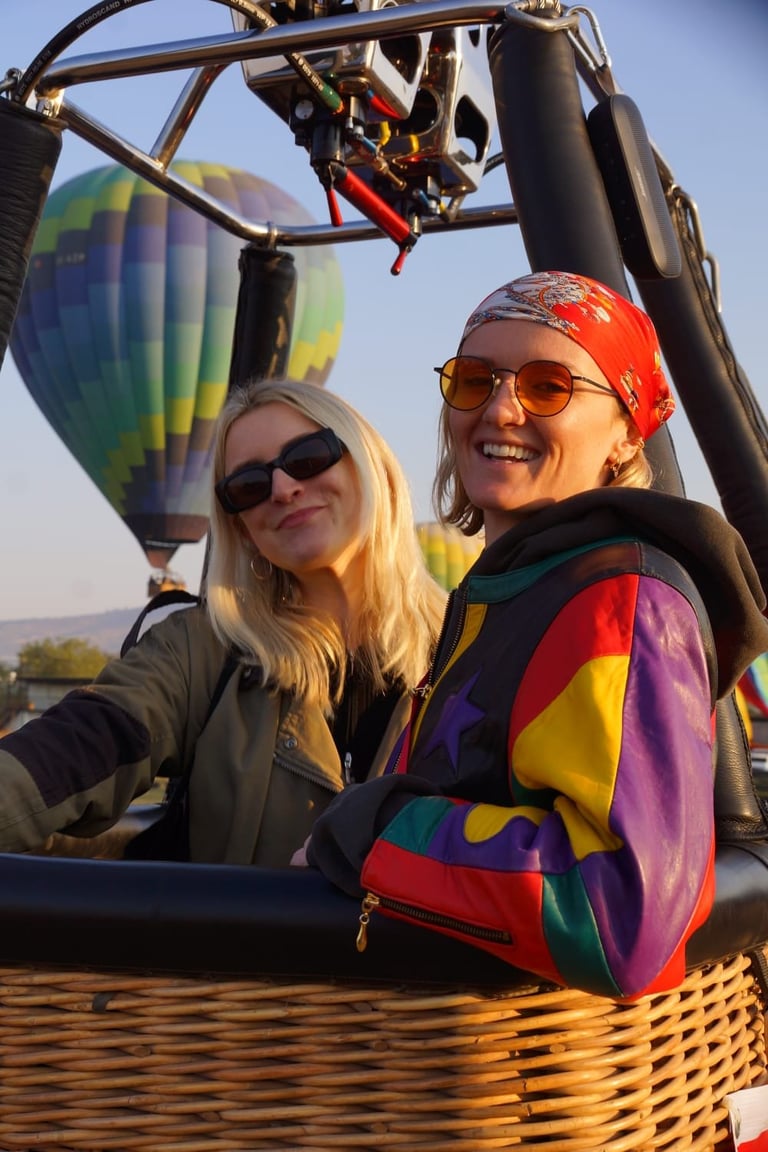 Two smiling women in a hot air balloon basket with colorful balloons visible in the background
